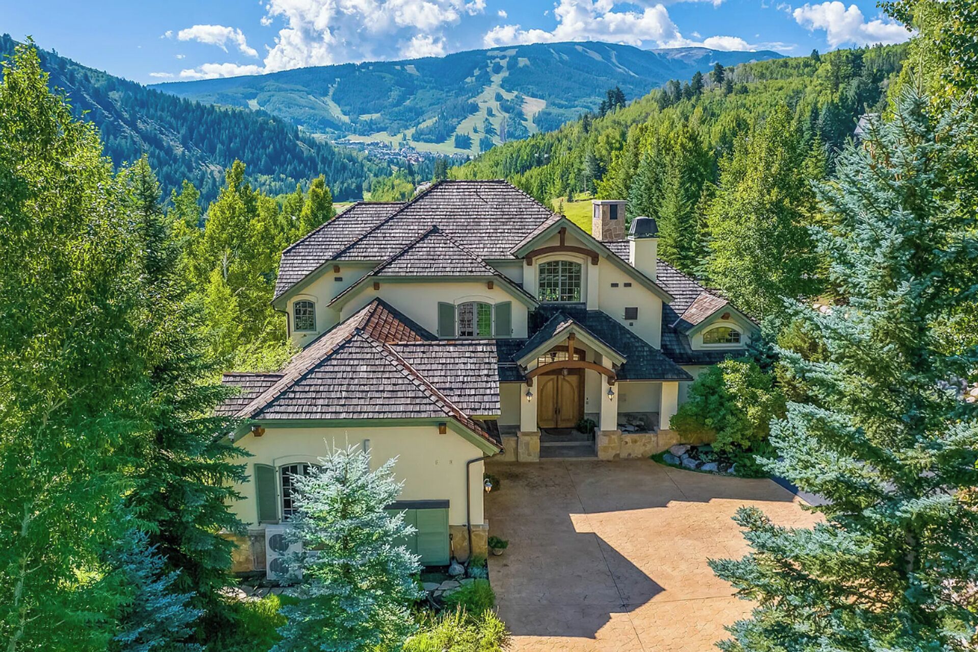 Una casa grande de dos pisos con un techo pronunciado, rodeada de un bosque denso y montañas al fondo.