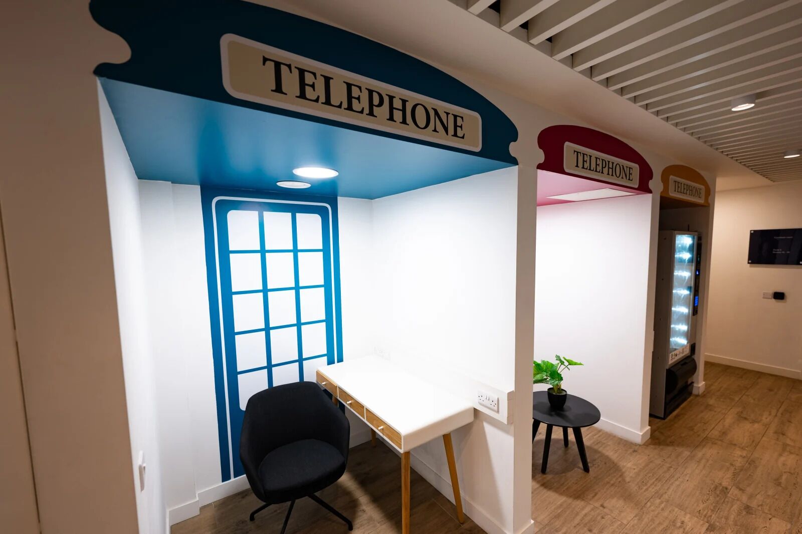A modern office space with a blue and white color scheme, featuring a desk and chair, a potted plant, and multiple telephone booths.