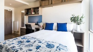 A modern hotel room with a large bed, a desk, and a chair. There is a radiator, a shelf with books, and a plant on the bedside table. - Student Only Arbury Court in Kingston (New Malden)