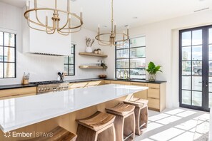 A modern kitchen with a large island, bar stools, pendant lighting, and a view through large windows. - The Cove at Desert Color (by Ember Stays) (St. George)