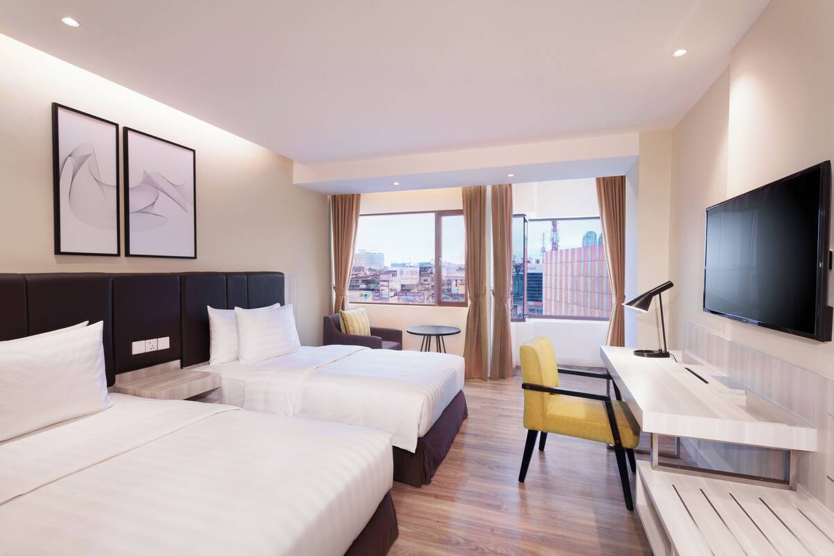 A hotel room with two beds, a desk, a chair, a TV, and a view of the city.
