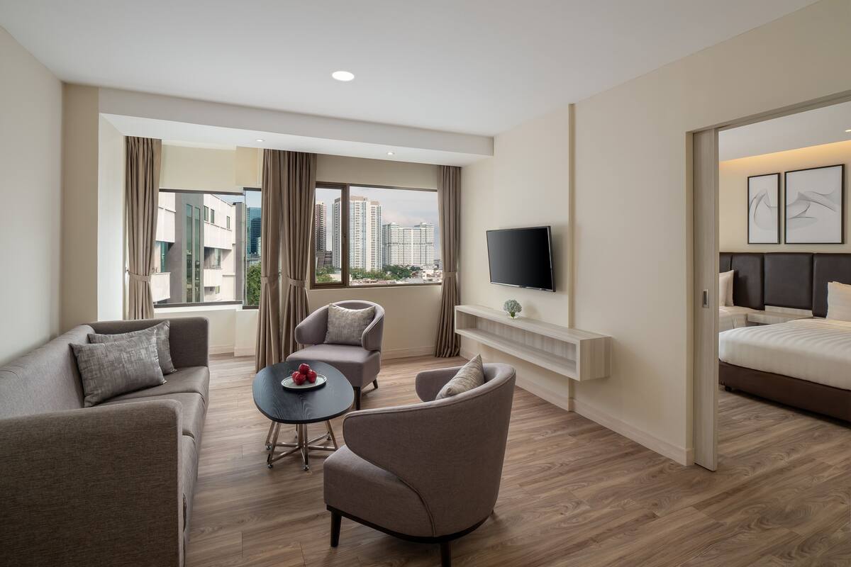 A modern hotel room with a sofa, armchair, and a small table with fruit. A TV is mounted on the wall, and there is a bed with a headboard.