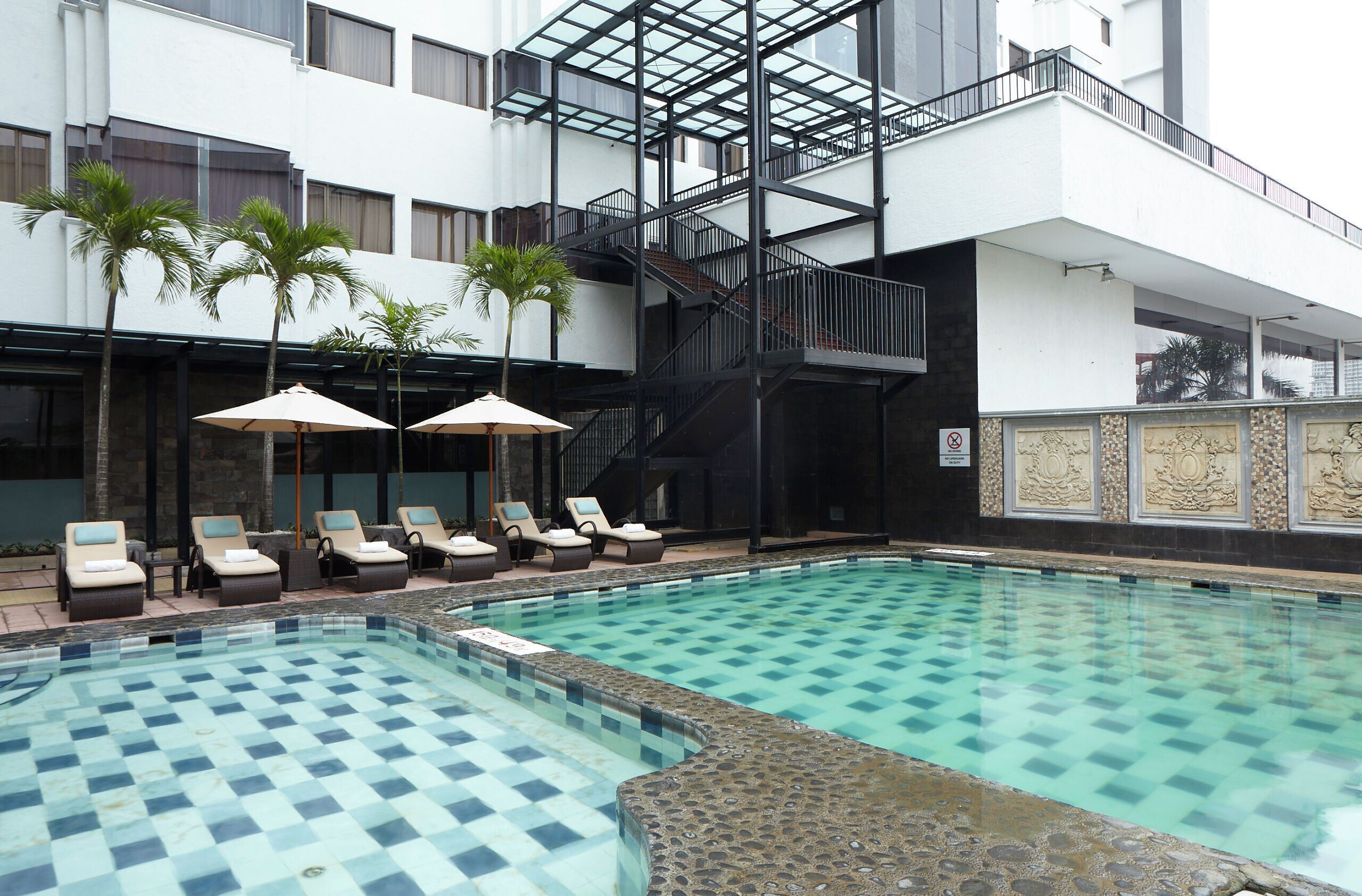 Un área de piscina con sillones de descanso y sombrillas, una escalera y la fachada de un edificio con paneles decorativos.