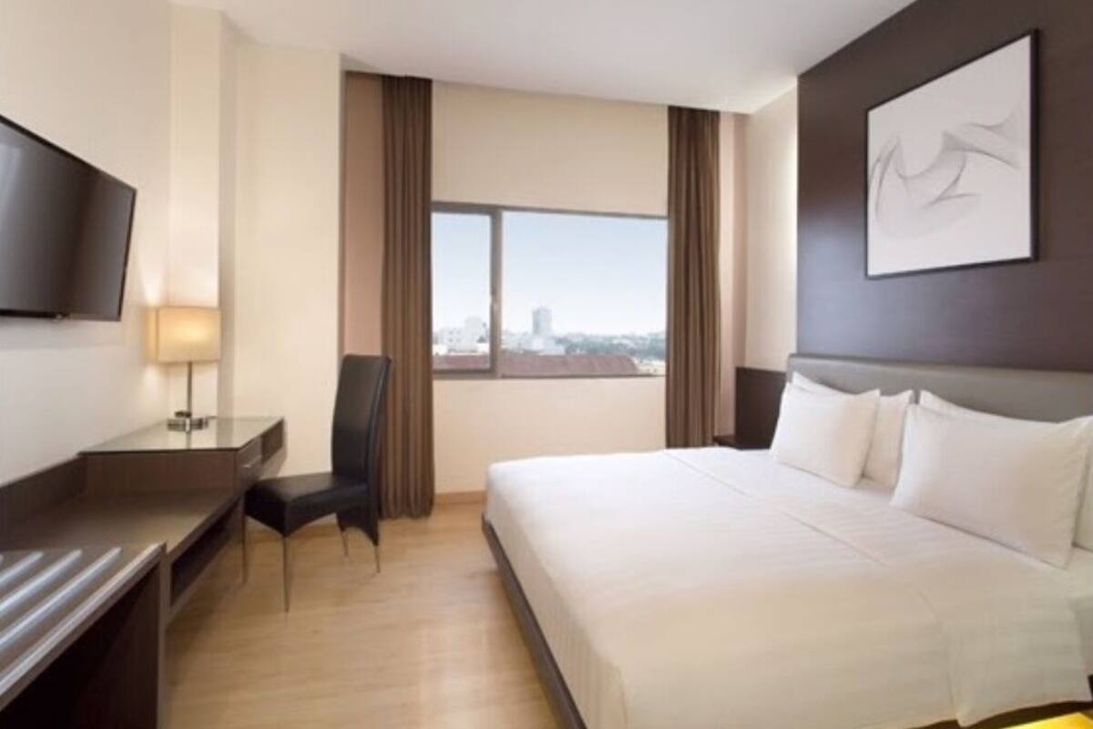 A hotel room with a large bed, a desk with a chair, a flat-screen TV, and a window with a view of the city.