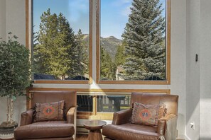 Una habitación con sillones de cuero, una mesita de madera y ventanas amplias con vistas a árboles y montañas.
