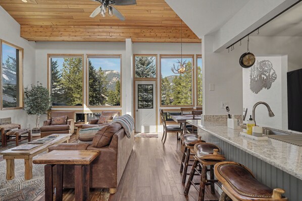 Amplia sala de estar con techo de madera, ventanas grandes que ofrecen vistas a árboles y montañas, zona de comedor con barra y cocina equipada con electrodomésticos modernos.