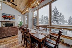 Comedor con mesa de madera dispuesta para cuatro personas, rodeada de sillas, con chimenea y un cuadro en la pared.