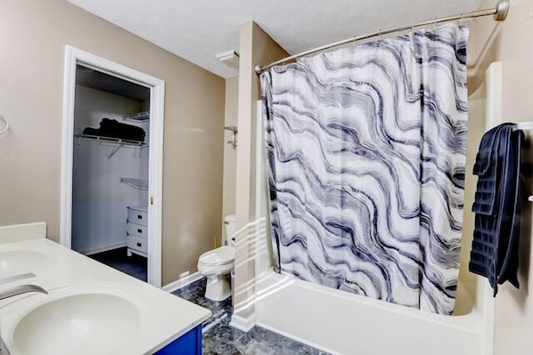 Un baño con lavamanos de mármol, cortina de ducha con diseño de mármol y vestidor con ropa colgada.