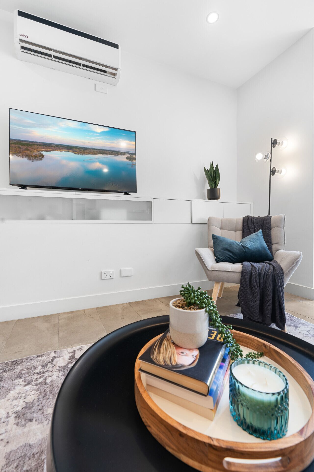 Uma sala de estar moderna com uma televisão de tela plana, um sofá com manta, um vaso de planta e uma bandeja com livros e uma vela.