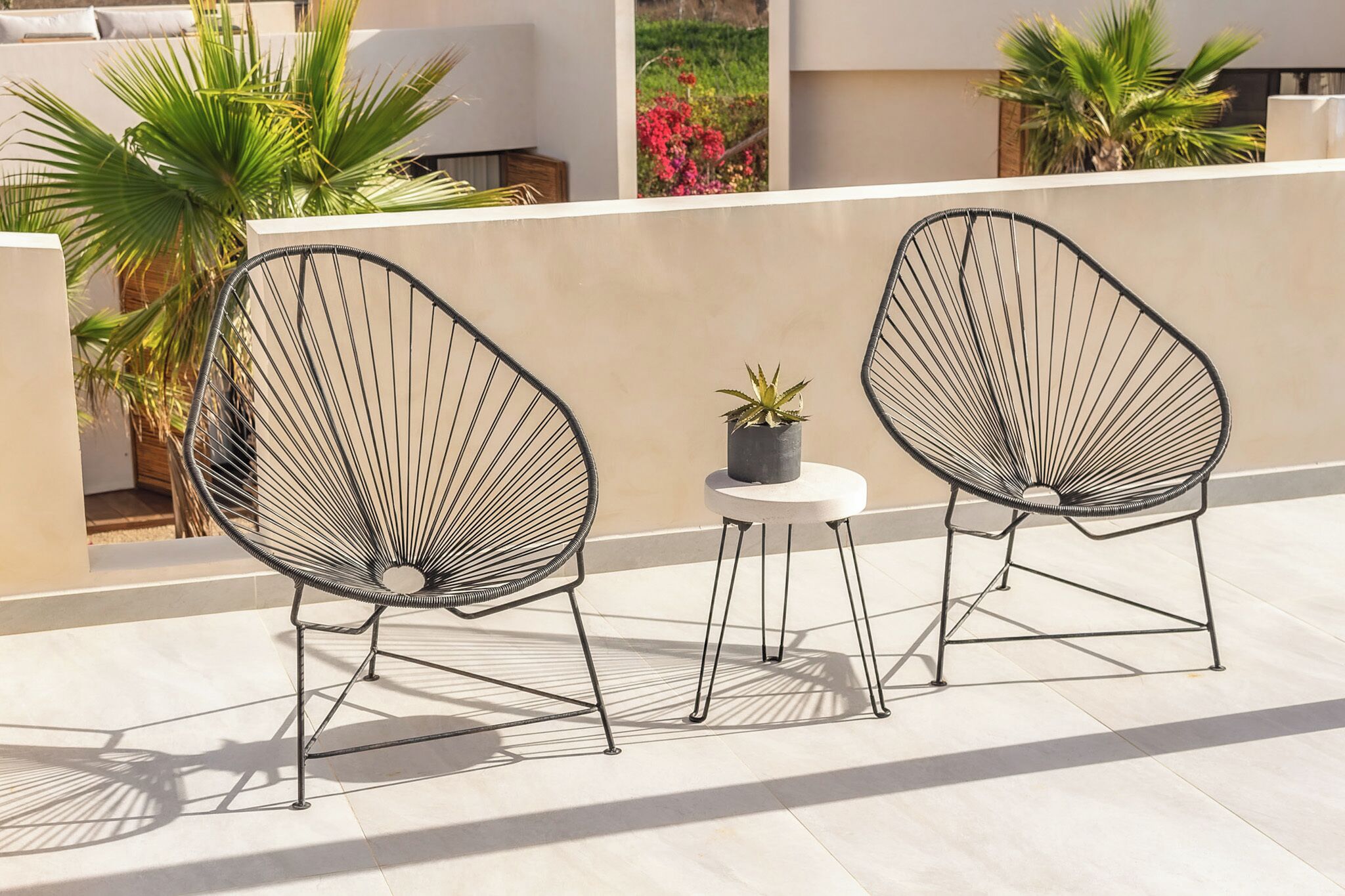 Two black wireframe chairs with a small table and a potted plant between them on a sunlit terrace with palm trees and flowers in the background.