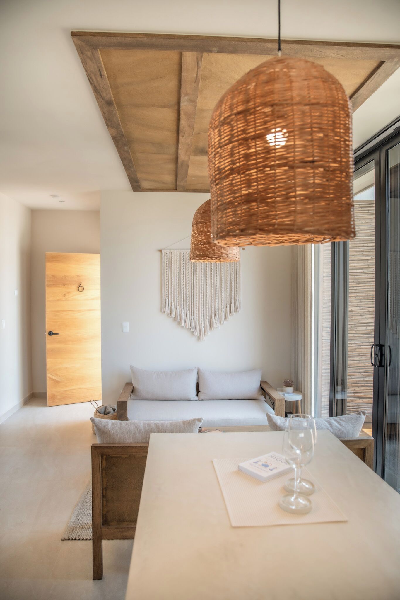 Ein modernes Zimmer mit Holzboden, einer großen gewebten Pendelleuchte, einem weißen Sofa, einem Holztisch mit einem Glas und einem Wandbehang aus Makramee.
