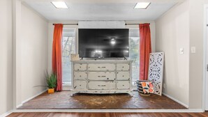 A room with a large flat-screen TV mounted on the wall, a white dresser with multiple drawers, a patterned rug on the floor, and a decorative panel on the wall. - 4 bedroom, King Bed, 4 min to Fairgrounds, TVs in all Rooms (Perry)