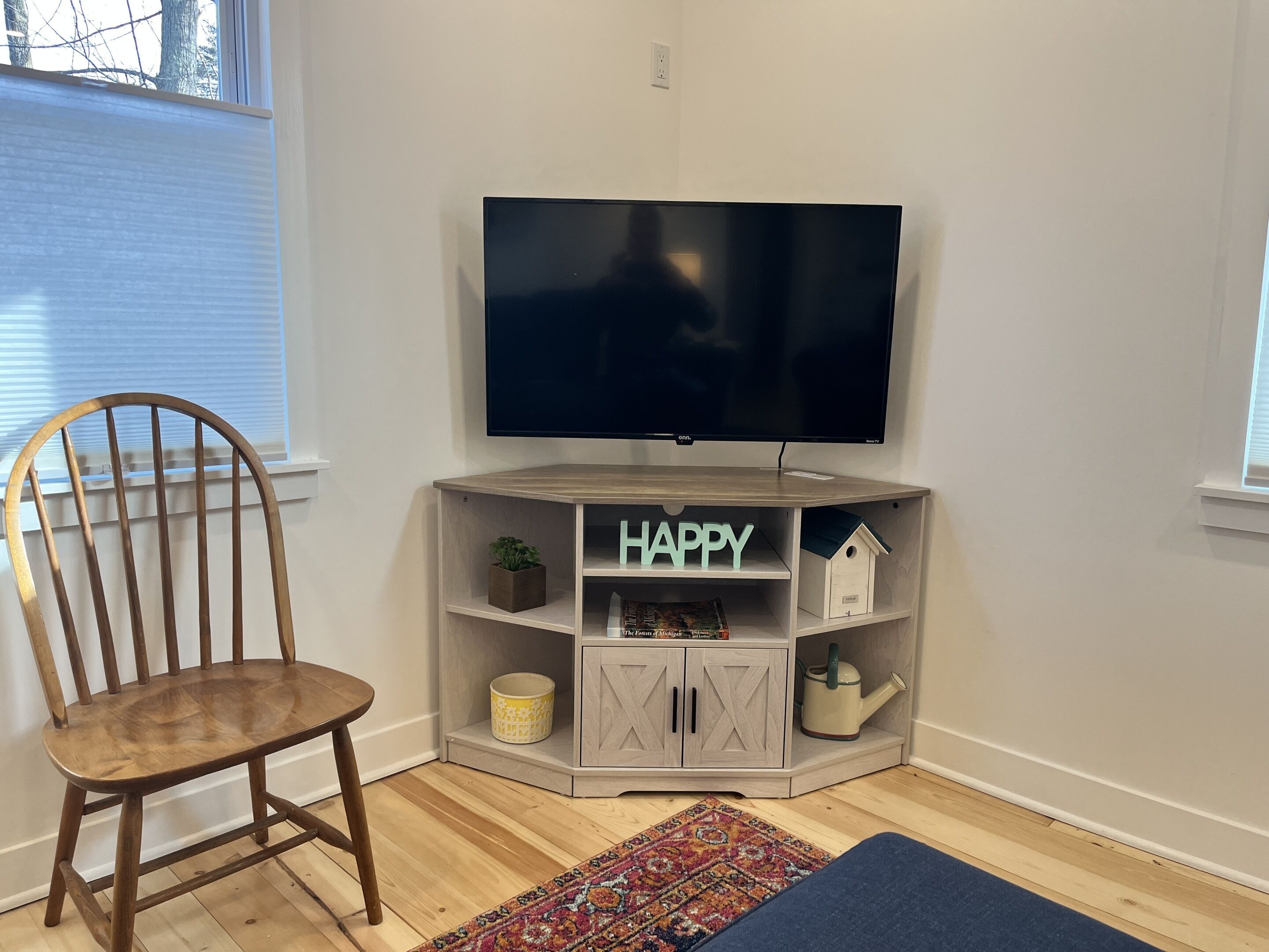 Ein Eckzimmer mit Fernseher auf einem Holzboden, einem Sessel, einem Regal mit der Aufschrift 'HAPPY' und einem kleinen Hausmodell.