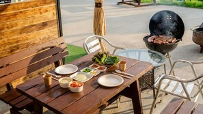 An outdoor dining area with a wooden table set for a meal, a grill in the background, and a glass table. - Yeosu Everyday Small Changes Pension (Yeosu)