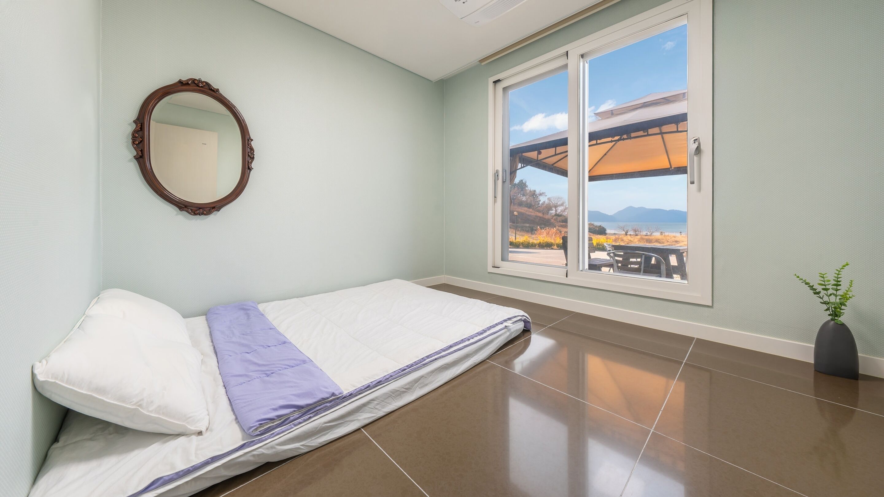 A room with a bed, a window with a view of the outdoors, and a mirror on the wall.