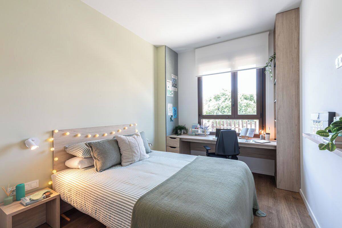Un dormitorio con cama, mesita de noche, escritorio, silla y ventana con vistas a árboles.