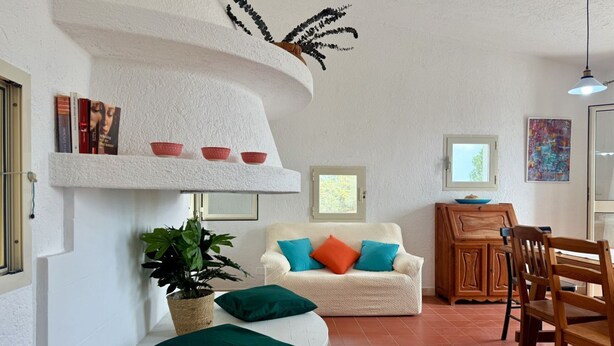 Una sala de estar acogedora con una pared blanca con textura, un sofá blanco con cojines coloridos, un aparador de madera, una mesa de comedor con sillas y una planta en maceta.