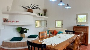 Un comedor con una mesa de madera, sillas y un mantel blanco. Hay plantas en macetas y una estantería con objetos decorativos.
