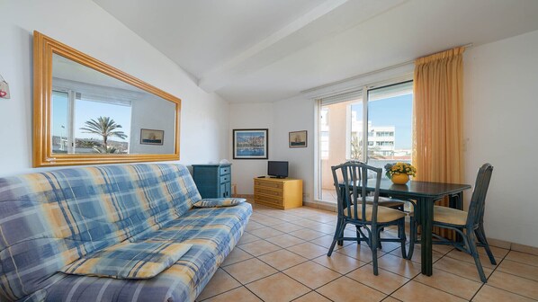 Una sala de estar con sofá, televisor, mesa con sillas y balcón con cortinas.