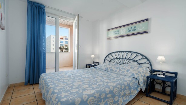 A bedroom with a bed, two bedside tables, a window with blue curtains, a view of buildings outside, and a sign on the wall. - Bright apt with large balcony for 4 pers (Agde)