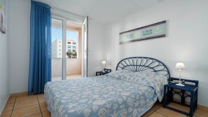 A bedroom with a bed, two bedside tables, a window with blue curtains, a view of buildings outside, and a sign on the wall.