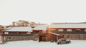 Un edificio de dos plantas con fachada de piedra y techo rojo, mostrando un terreno cubierto de nieve y un vehículo estacionado al frente.