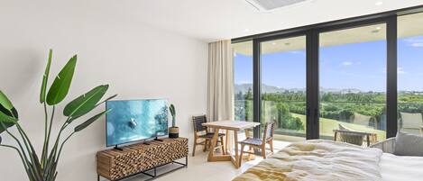 A modern hotel room with a large bed, a TV, a dining table, and a view of a lush landscape.