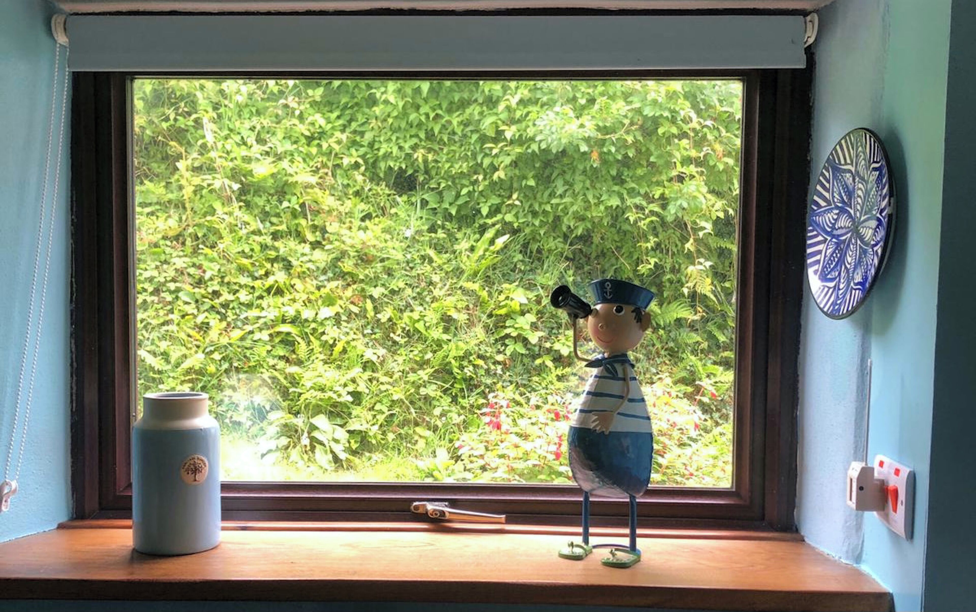A window with a view of greenery, a small figurine, a ceramic pot, and a decorative plate.