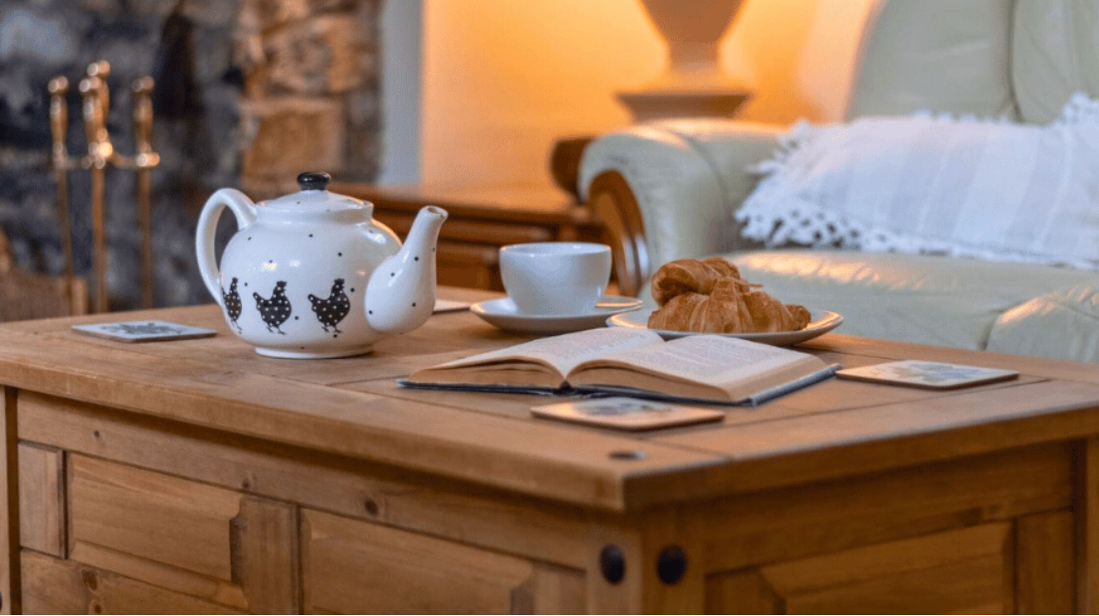 Une table en bois avec une théière, une tasse, une assiette de croissants et un livre ouvert.