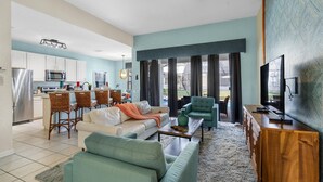 Un salon moderne comprenant un canapé, des fauteuils, une table basse et un téléviseur. Il y a également un coin cuisine avec des placards et des appareils électroménagers.