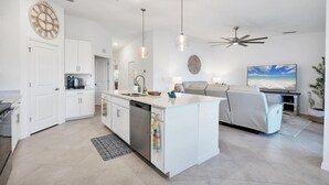 Une cuisine moderne avec des meubles blancs, un îlot central, des luminaires suspendus et un ventilateur de plafond.