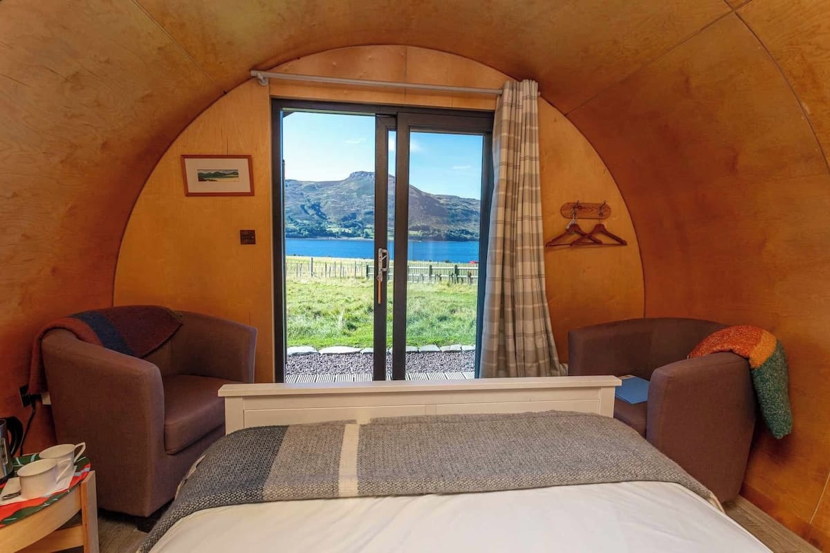 Habitació acollidora amb un llit, dues butaques i una finestra gran que ofereix una vista panoràmica d'un llac i muntanyes.