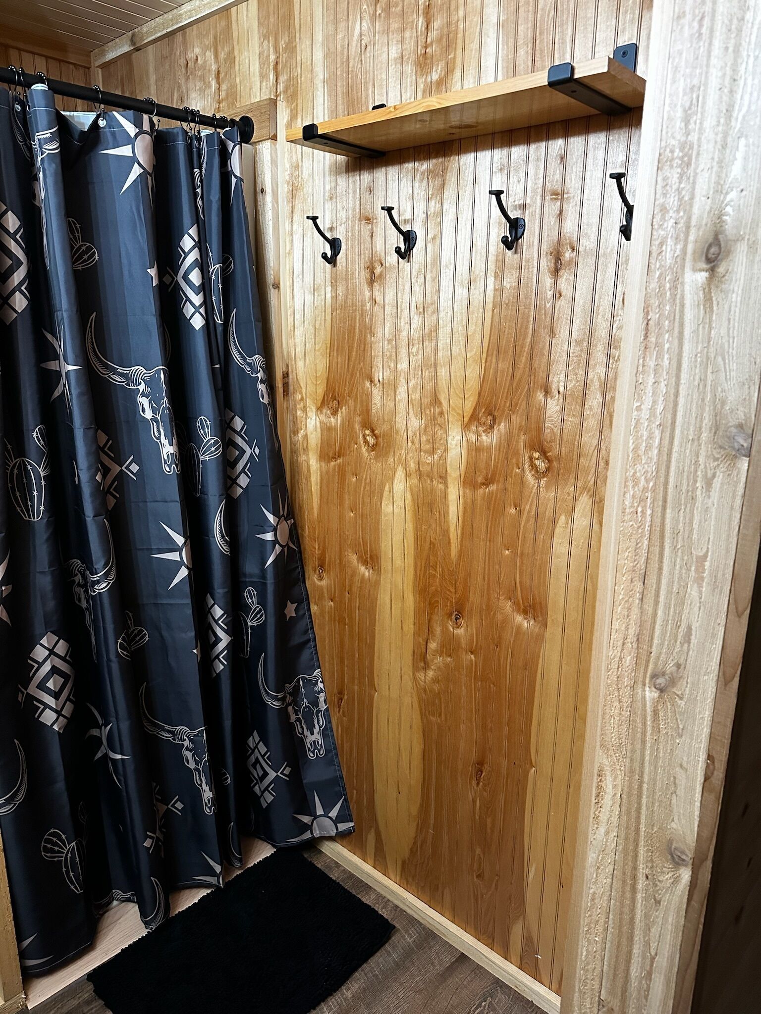 A bathroom with a wooden wall, a shower curtain with a geometric pattern, and a black rug on the floor.