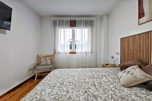 Un dormitori amb un llit, una cadira, una tauleta de nit, una finestra amb cortines translúcides i un capçal de llit de fusta.