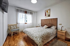 A bedroom with a wooden floor, a bed with a patterned bedspread, a wooden nightstand, a chair, and a window with sheer curtains.