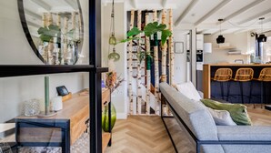Een moderne woonkamer met een bank, een eethoek met stoelen, een keuken op de achtergrond en een grote spiegel aan de muur.