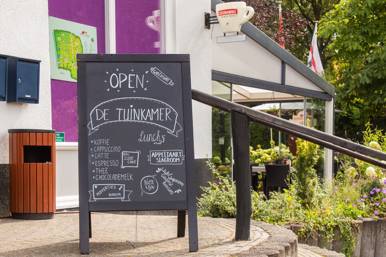 A chalkboard sign outside a building advertising 'Open' hours and 'De Tuinkamer' with a list of food and drink options.