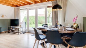 Un coin repas avec une table en bois dressée pour quatre personnes, entourée de chaises noires, d’un téléviseur et d’une grande fenêtre agrémentée de rideaux.