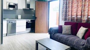 Een moderne keuken met een magnetron, oven en vaatwasser. Een houten deur leidt naar een balkon. In de woonkamer staat een bank met gestreepte kussens.