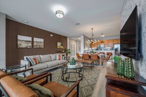 Una sala de estar moderna con sofá, mesa de centro y sillones. Se observa una zona de cocina con armarios de madera y una mesa de comedor.