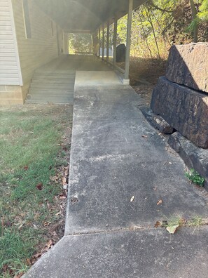 A concrete pathway leading to a covered porch area with a stone bench and a grill.