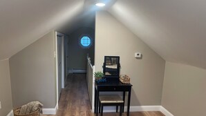 Um quarto com teto inclinado, piso de madeira, uma mesa pequena com um espelho, um cesto e uma cadeira.