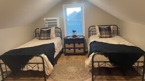 Um quarto de sótão pequeno e triangular com duas camas de solteiro, uma mesa pequena e uma janela com persianas.