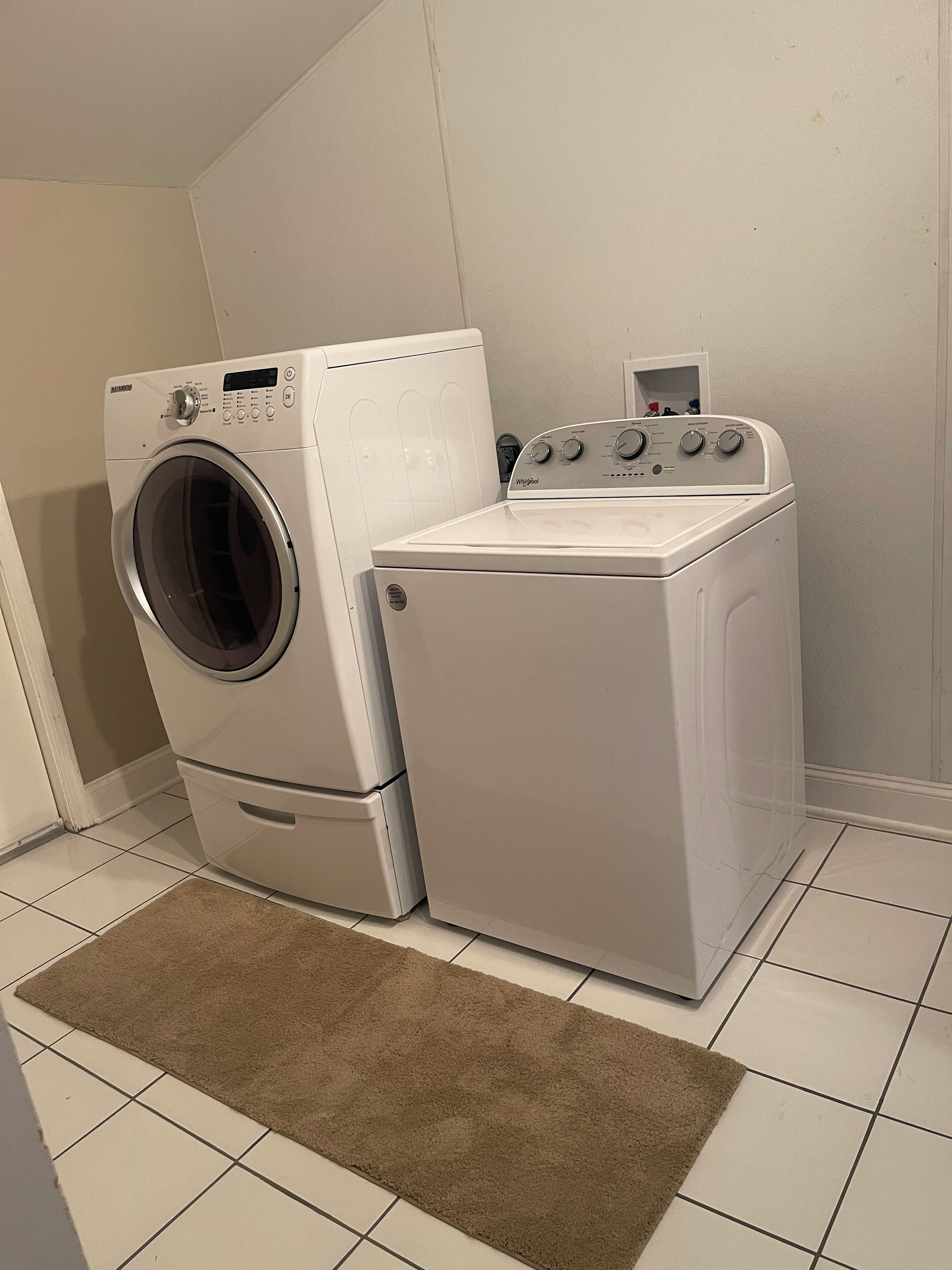 A laundry room with a front-loading washer and dryer, a brown rug, and white tiled flooring.