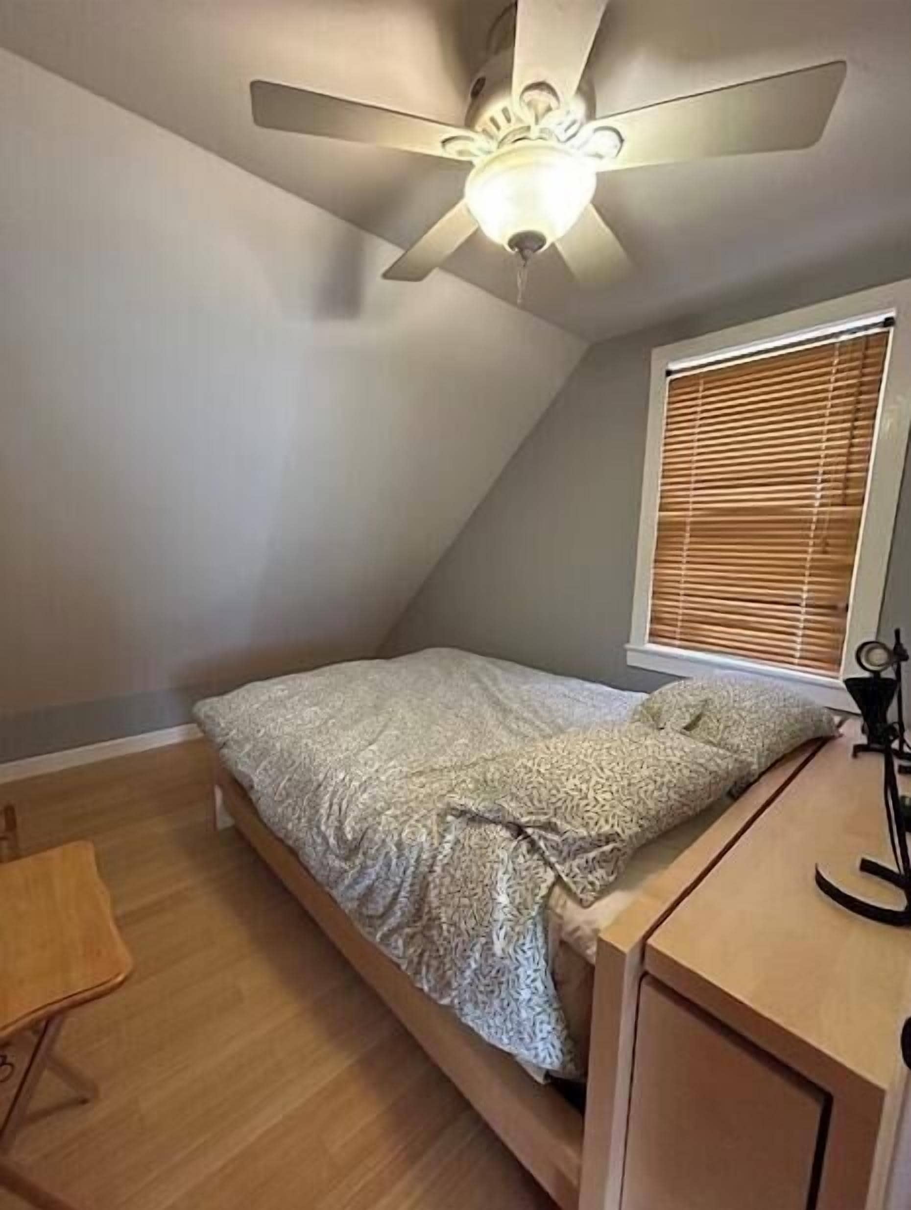 Ein kleines Zimmer mit einem Einzelbett, geneigter Decke und Deckenventilator. Es gibt ein Fenster mit Jalousien, einen Nachttisch und einen kleinen Schreibtisch mit Stuhl.