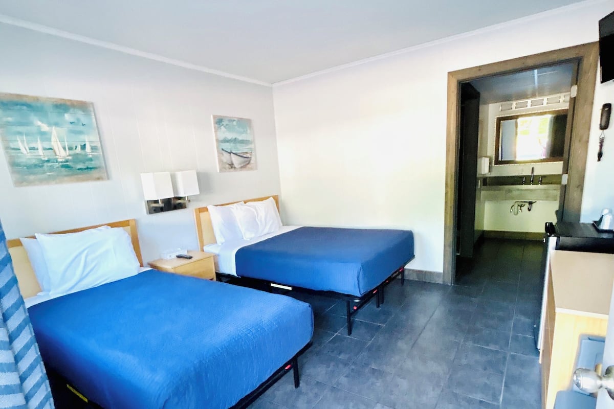 A hotel room with two beds, a small desk, a mirror, and a bathroom visible through an open door.