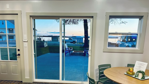 A room with a sliding glass door leading to an outdoor area, a round wooden table with chairs, and a view of a blue building through the door. - Silver Sands Hotel & Resort (Mears)