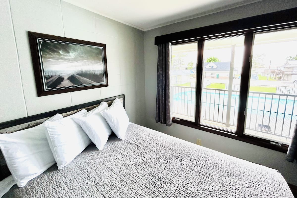A hotel room with a bed, a framed picture on the wall, and a window with a view of a swimming pool and buildings.