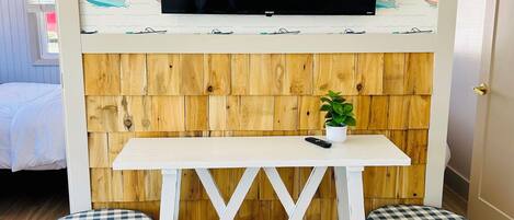 Una habitación con un televisor montado en una pared con motivos náuticos, un panel de pared de madera, una mesa blanca con dos otomanas a cuadros y una pequeña planta en maceta.
