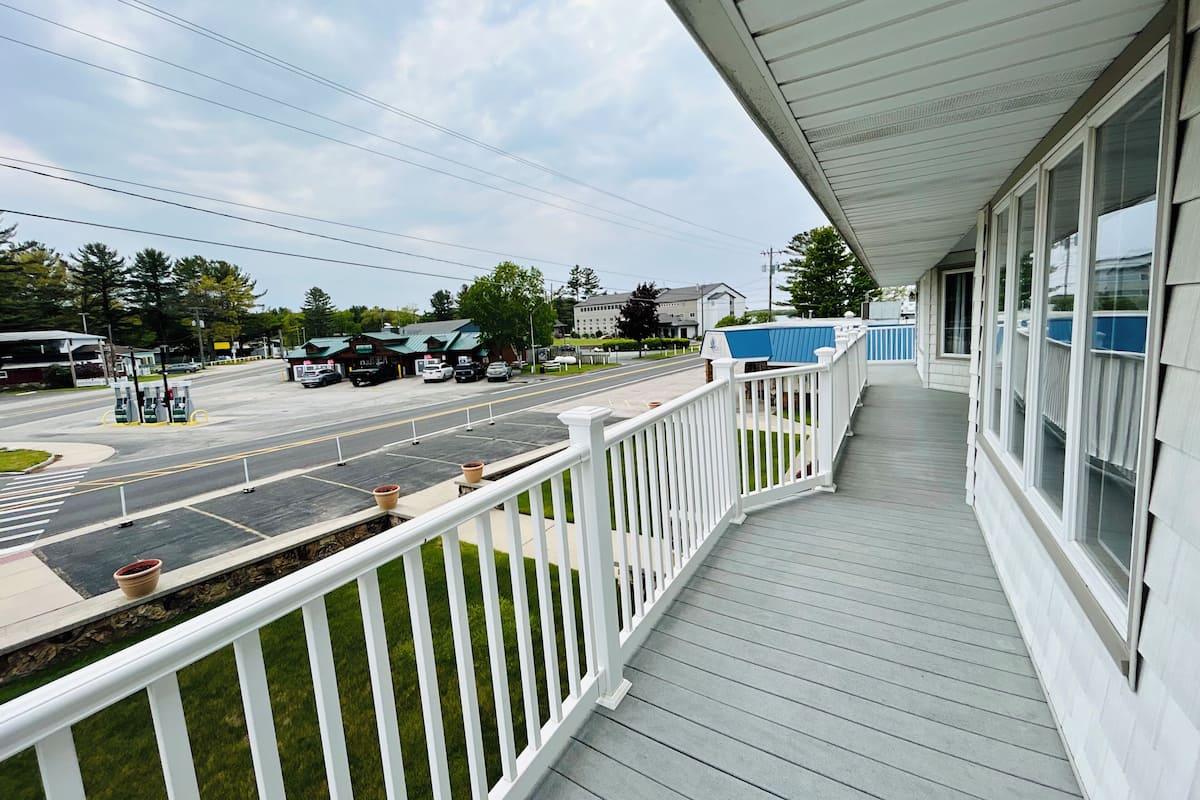 A covered porch with a white railing, a view of a street with parked cars, and a building with a green roof in the background.